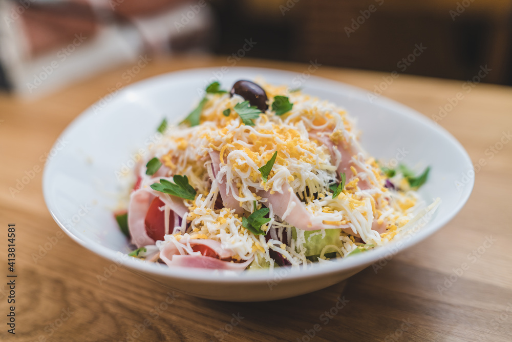 Close shot of shopska salad egg,bacon, arugula,lettuce salad in white bowl on brown wooden table with olive on top Close shot of shopska salad egg,bacon, arugula,lettuce salad in white bowl on brown wooden table with olive on top