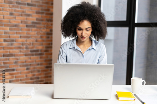 Diligent young American businesswoman sitting at the laptop, typing a new work strategy, creating a new startup, writing notes, organizing the conference, chatting with olleagues photo