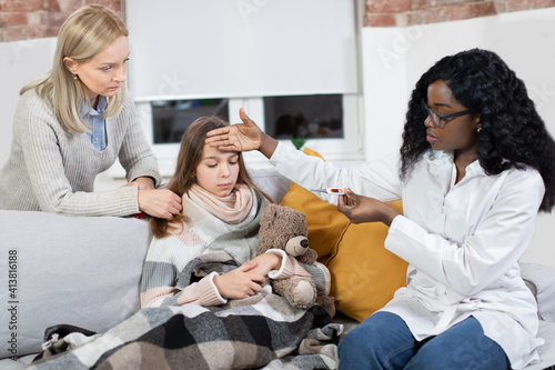 Home treatment under doctor s supervision. Young professional female black doctor making home visit  checking temperature of her patient  sick teen girl  supporting by mom