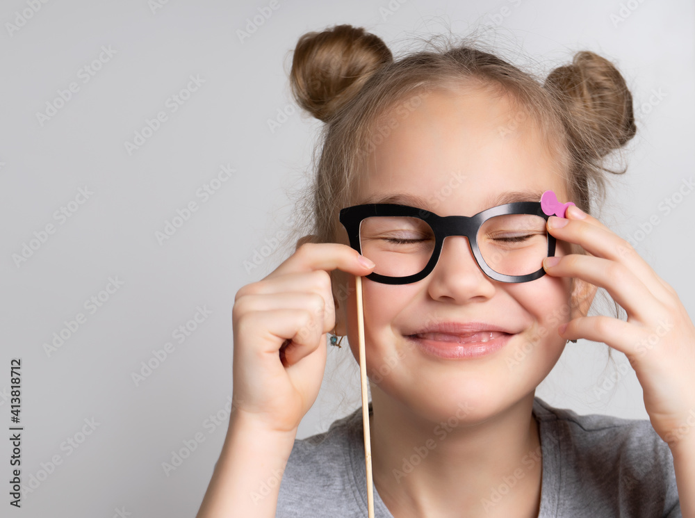Happy girl with eye closed wearing paper glasses mask studio portrait shot. Photo booth accessory for children birthday party celebration. Glad caucasian female child headshot on grey background