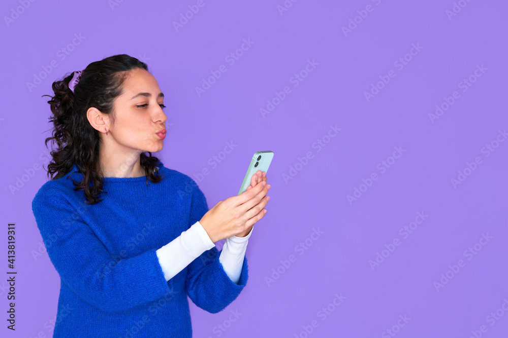 Pretty young woman in blue sweater posing isolated on purple background. People lifestyle concept. Copy space. Doing selfie shot on mobile phone, blowing and sending air kiss Pretty young woman in blue sweater posing isolated on purple background. People lifestyle concept. Copy space. Doing selfie shot on mobile phone, blowing and sending air kiss