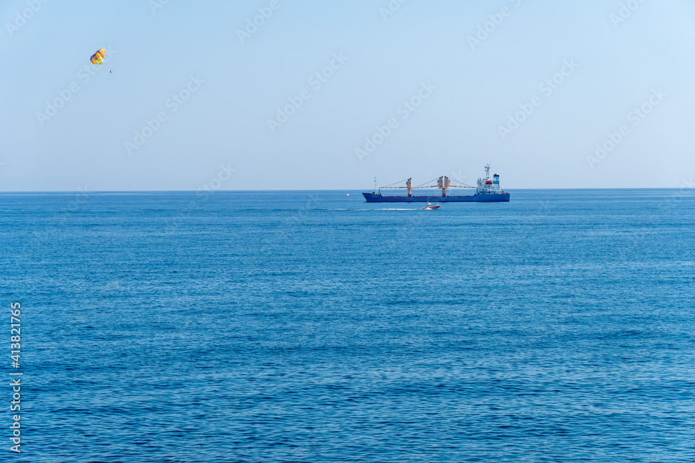 Colored para sail wing pulled by a boat in the sea water, Parasailing also known as parascending or para kiting. Container ship in a background, negative space. Colored para sail wing pulled by a boat in the sea water, Parasailing also known as parascending or para kiting. Container ship in a background, negative space.