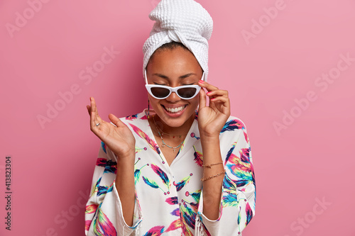 Positive happy woman with dark skin wears sunglasses dressing gown and wrapped towel on head keeps hand raised has fun on domestic party isolated over pink background. People and style concept © wayhome.studio 