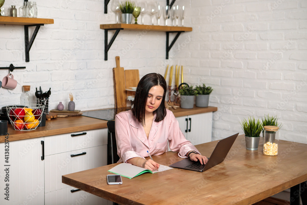 A woman works in the office remotely from home in the kitchen, sitting at the computer. Distance learning online education and work. Girl student prepares for exams. A woman works in the office remotely from home in the kitchen, sitting at the computer. Distance learning online education and work. Girl student prepares for exams.