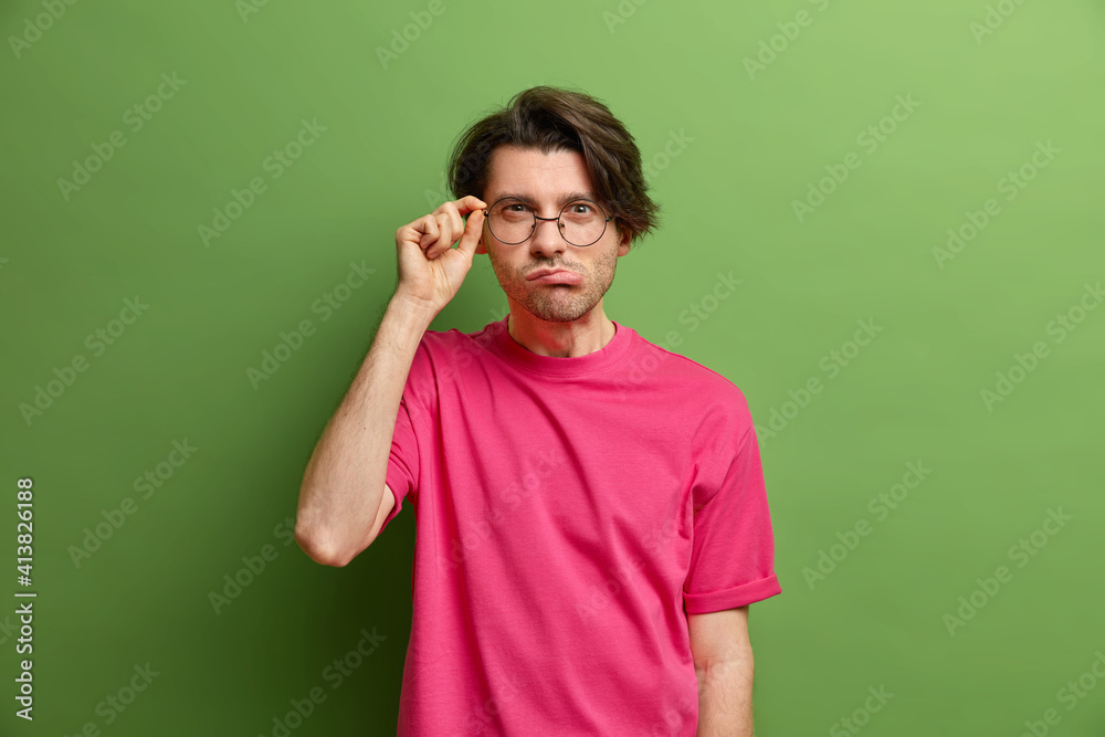 Horizontal shot of serious guy looks attentively through spectacles at camera wears casual pink t shirt has mysterious gaze purses lips poses over green background tries to see something in distance