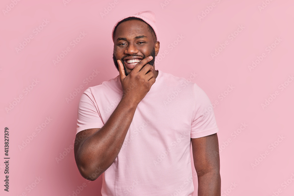 Portrait of handsome bearded man holds chin smiles broadly being in good mood wears hat and casual t shirt isolated over pink background. Happy African American male model hears someting funny