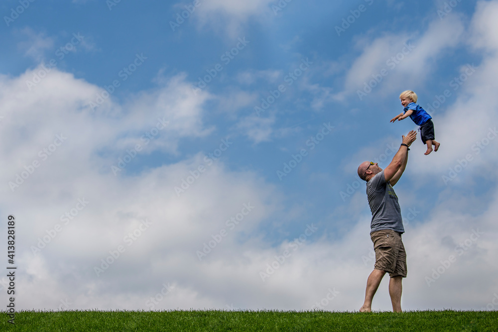 Side view of playful father throwing son in mid-air while standing on grassy field against cloudy sky Side view of playful father throwing son in mid-air while standing on grassy field against cloudy sky