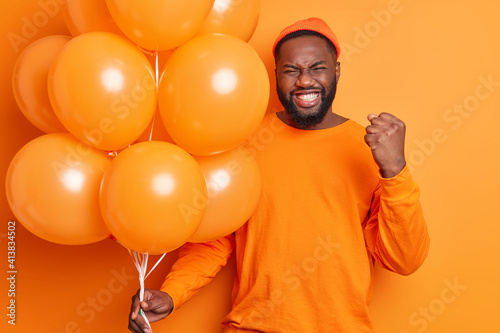 Happy emotional dark skinned bearded guy clenches fist and teeth celebrates getting promotion spends free time at party dressed casually holds inflated balloons isolated over orange background © wayhome.studio 