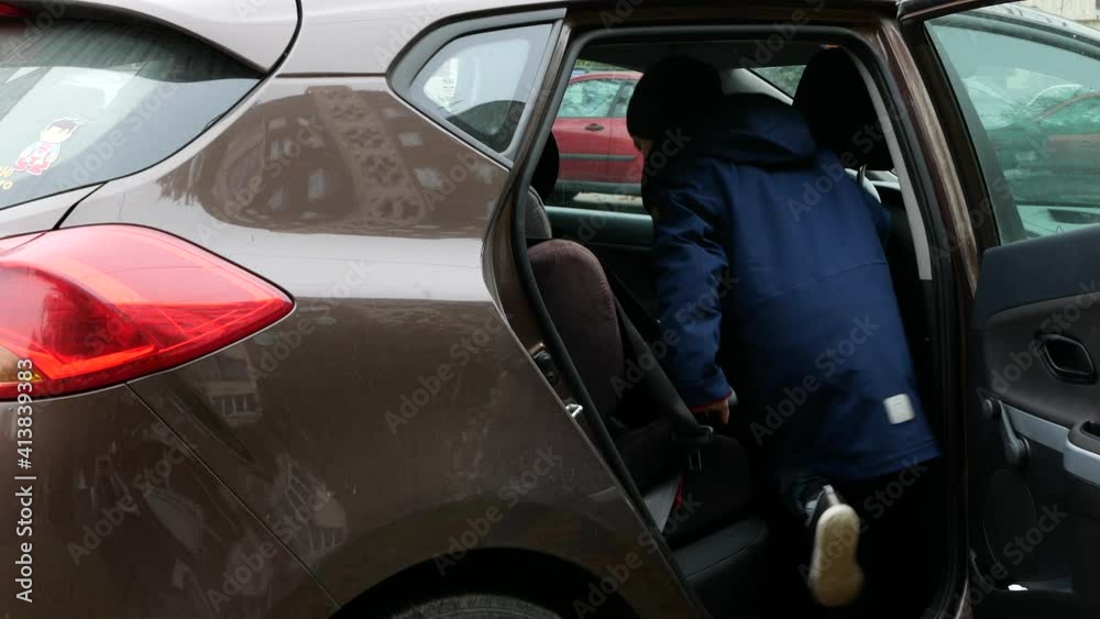 Woman opening the door and happy little boy in navy blue anorak getting into a car to go on a journey