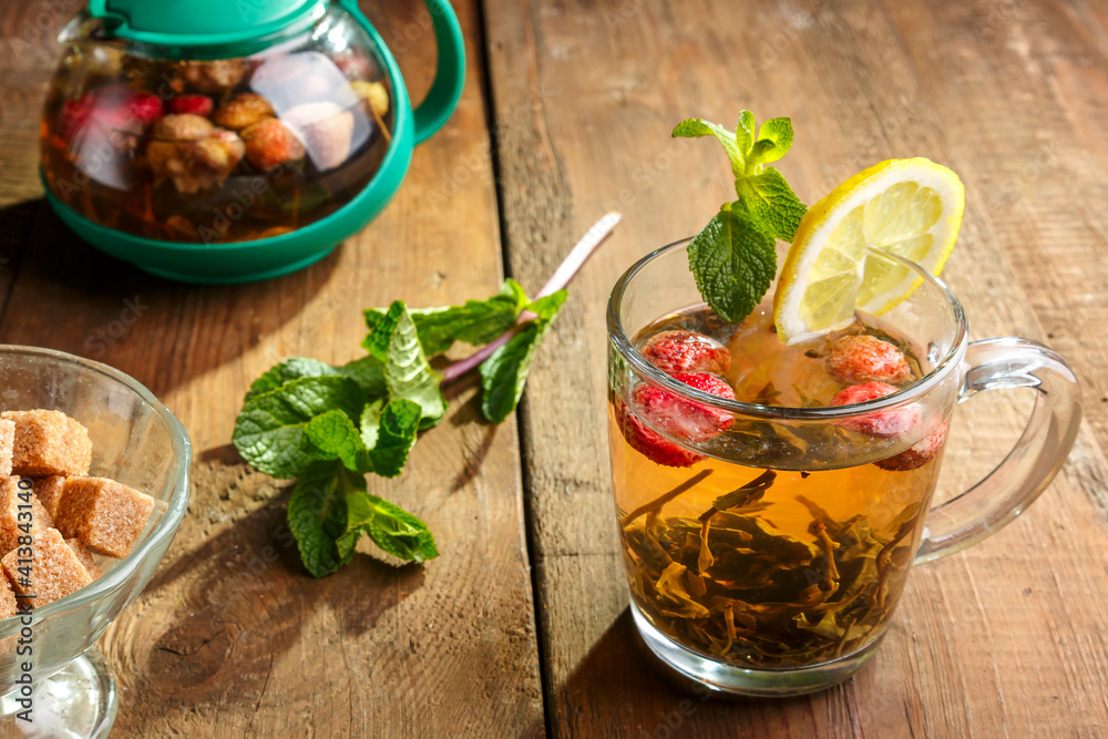 green tea in a glass cup with strawberries mint and lemon on a wooden table and a teapot and mint leaves.