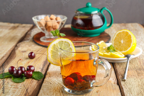 black tea in a glass cup with mint cherries and lemon on a wooden table next to fresh cherries and a teapot and lemon in a plate.