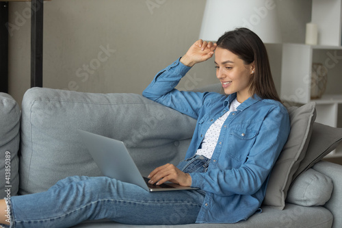 Having pleasure. Smiling teenage female rest on sofa with laptop on knees look at screen engaged in virtual chat online read good news. Peaceful young lady enjoy weekend at home spend time in internet