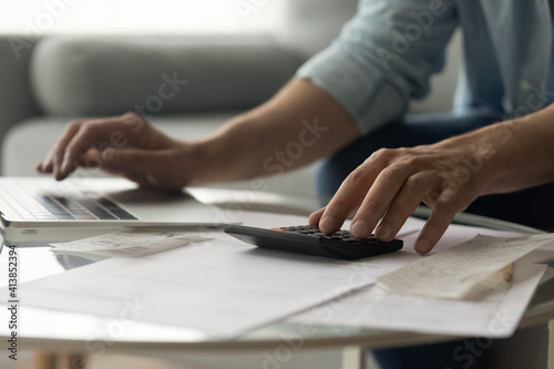 Close up of young male accountant hands make audit of expenses calculate charges based on paper documents. Cropped shot of man bookkeeper work with bills pay fees taxes online using web app on laptop photo