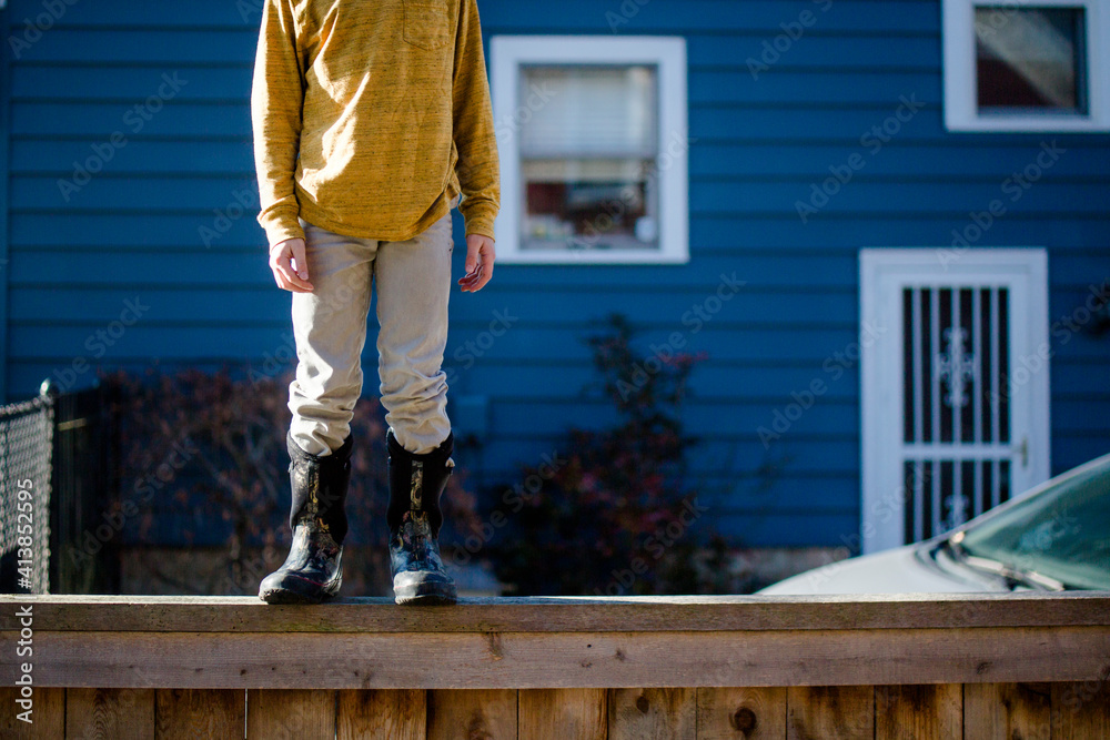 A child in boots and yellow shirt stands on fence against blue house A child in boots and yellow shirt stands on fence against blue house
