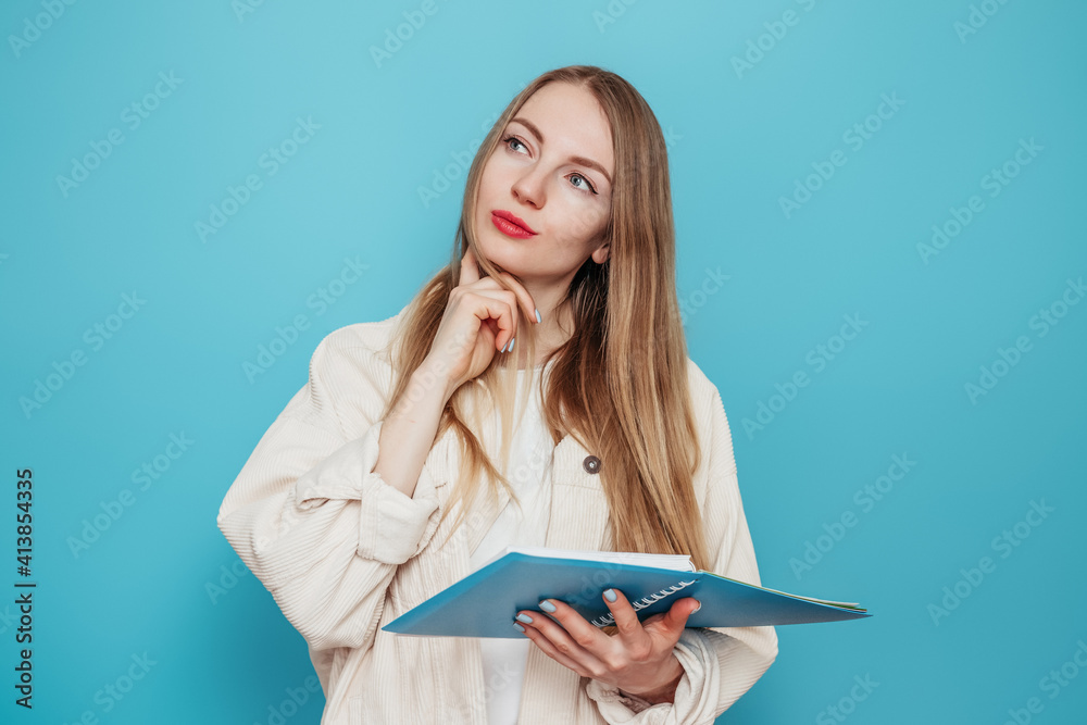 Pensive blonde student girl holding an open exercise book and looking to copy space isolated on blue background in studio. Pensive blonde student girl holding an open exercise book and looking to copy space isolated on blue background in studio.