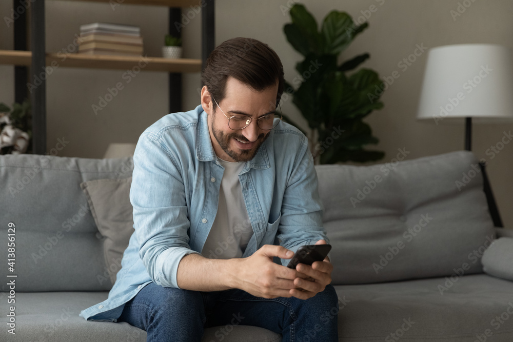 Positive millennial male holding phone smiling when reading funny post in social network blog chatting dating online. Young guy sit on sofa enjoy surfing web correspond with good friend in mobile app