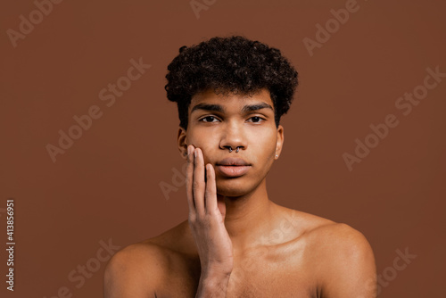 Photo of attractive black man with piercing touches his smooth face and looks at camera. Naked torso, isolated brown color background