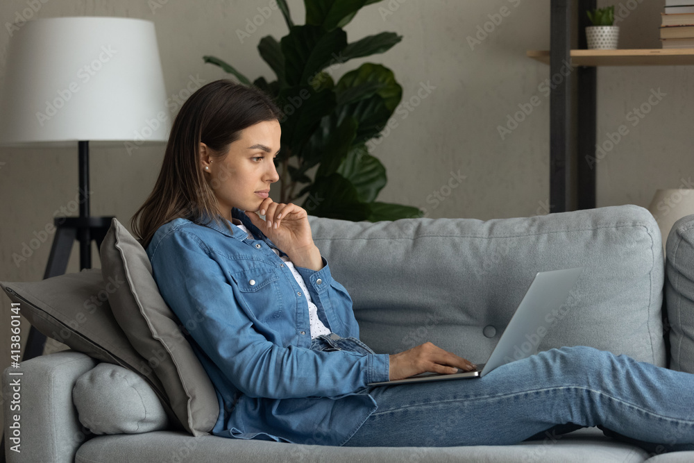 Pensive young woman student read post at web blog on laptop screen think on information received from internet. Concentrated teen female sit on couch write resume covering letter to potential employer Pensive young woman student read post at web blog on laptop screen think on information received from internet. Concentrated teen female sit on couch write resume covering letter to potential employer