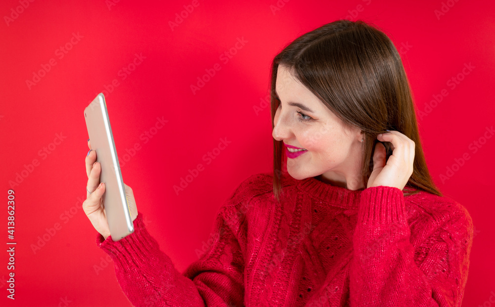 Naklejka premium Portrait of stunning joyful young woman in red fur sweater using tablet pc computer isolated on bright red wall background in studio