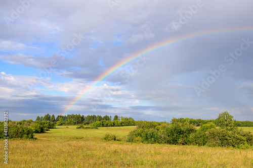 green field and parts of trees during the day with cumulus clouds and a rainbow in the sky