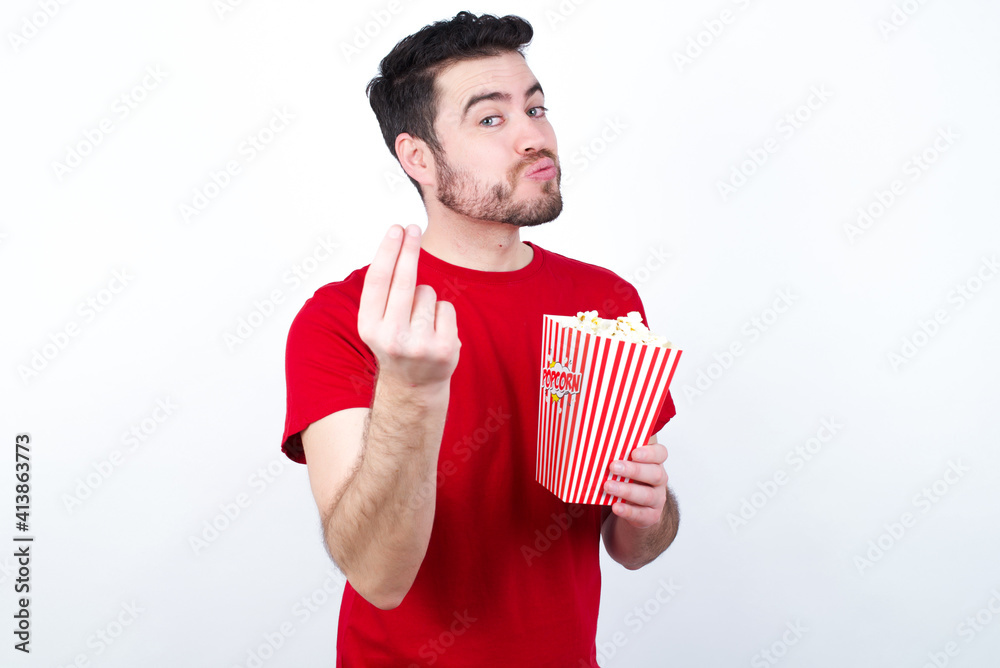 Young handsome man in red T-shirt against white background eating popcorn doing money gesture with hands, asking for salary payment, millionaire business Young handsome man in red T-shirt against white background eating popcorn doing money gesture with hands, asking for salary payment, millionaire business