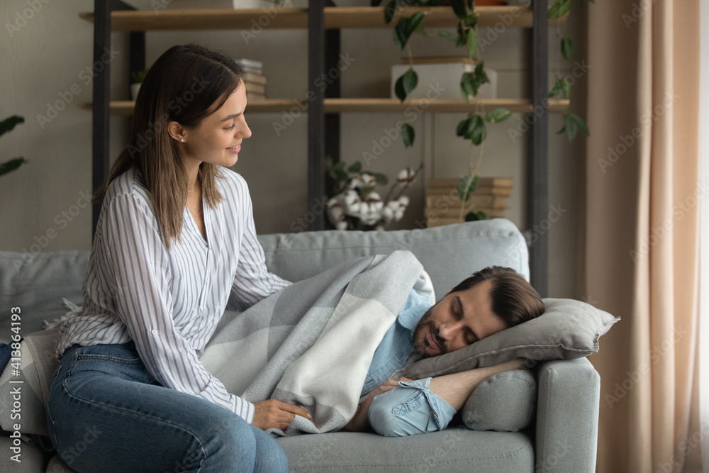 Obraz premium Love in details. Attentive millennial wife carefully covering tired husband sleeping on couch with warm soft plaid blanket. Affectionate young female take care of fatigued spouse resting on cozy sofa