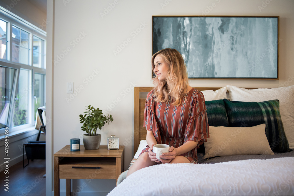 Young woman sitting on bed in apartment with coffee mug Young woman sitting on bed in apartment with coffee mug