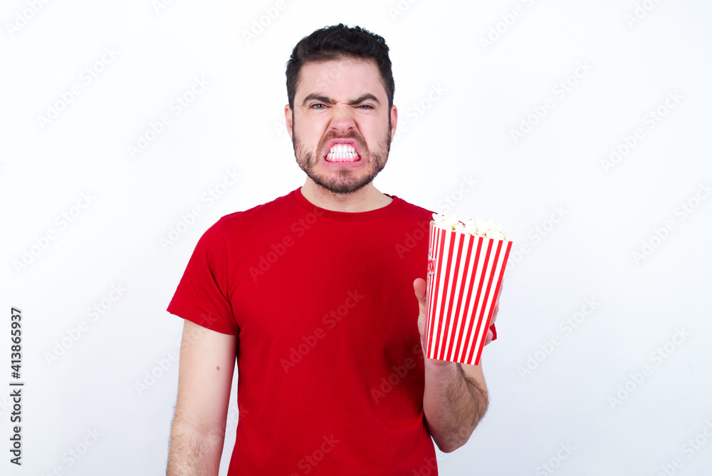 Young handsome man in red T-shirt against white background eating popcorn keeps teeth clenched, frowns face in dissatisfaction, irritated because of much duties. Young handsome man in red T-shirt against white background eating popcorn keeps teeth clenched, frowns face in dissatisfaction, irritated because of much duties.