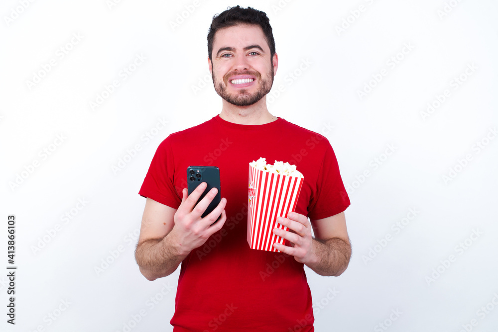 Young handsome man in red T-shirt against white background eating popcorn enjoys distant communication, uses mobile phone, surfs fast unlimited internet, has pleasant smile, makes shopping online, Young handsome man in red T-shirt against white background eating popcorn enjoys distant communication, uses mobile phone, surfs fast unlimited internet, has pleasant smile, makes shopping online,