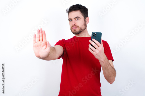 Young handsome man in red T-shirt against white background eating popcorn using and texting with smartphone with open hand doing stop sign with serious and confident expression, defense gesture