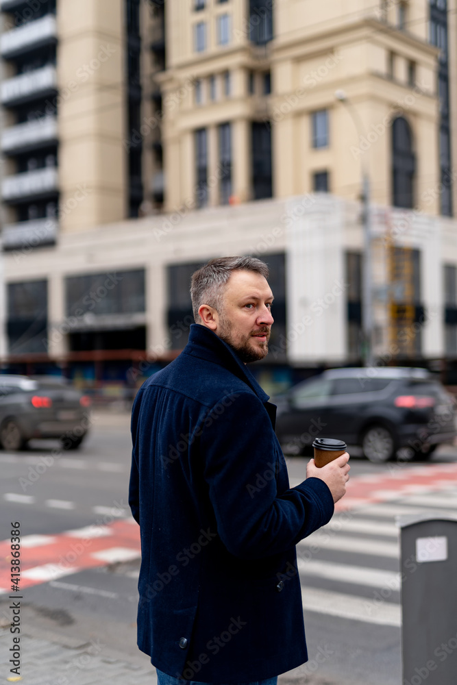 Male crosses the street in dark coat in the morning. Bearded man with morning coffee in hands. Closeup. Male crosses the street in dark coat in the morning. Bearded man with morning coffee in hands. Closeup.
