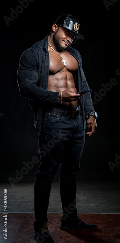 Strong athlete in dark jeans poses to the camera on black background. Unbuttoned black shirt and perfect abs. Portrait of an afro american bodybuilder with phone in hands.