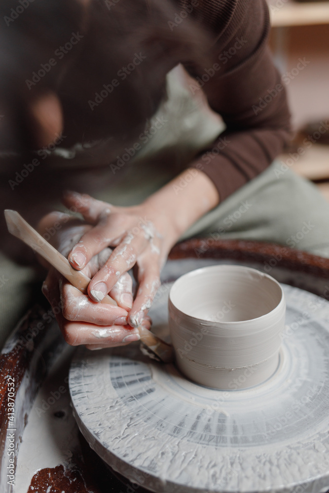 Closeup of potter's hands molds clay pot spinning on pottery wheel with special wooden tool. Woman works with tools in pottery workshopCloseup of potter hands molds clay pot spinning on pottery whee