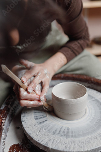 Closeup of potter's hands molds clay pot spinning on pottery wheel with special wooden tool. Woman works with tools in pottery workshopCloseup of potter hands molds clay pot spinning on pottery whee