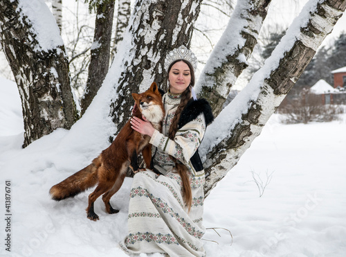 Russian beauty in a national costume with a red fox in a winter snowy forest 