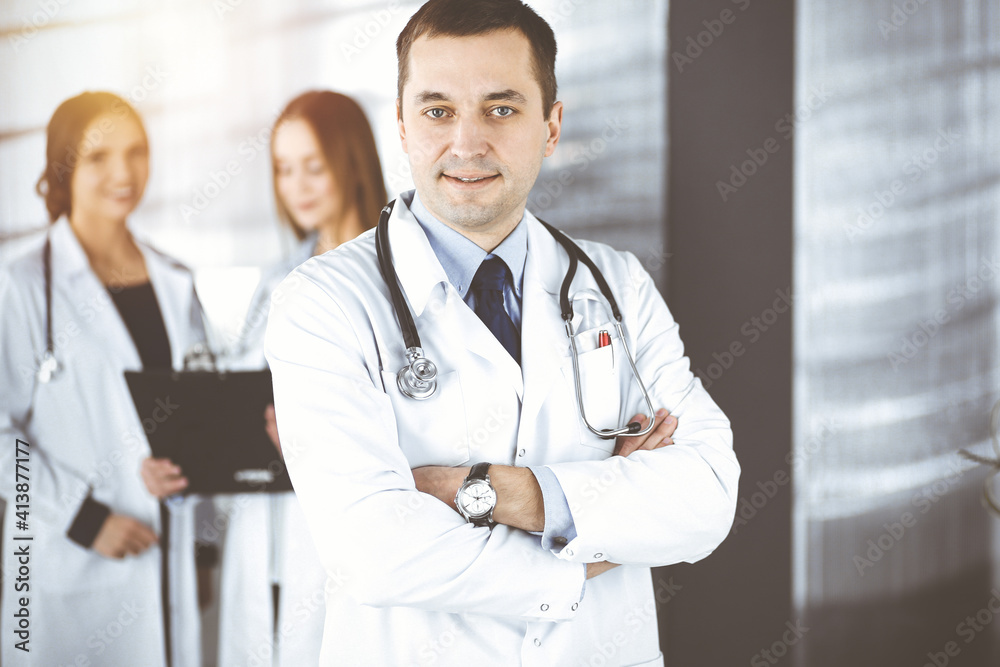 Professional middle aged doctor with a stethoscope and crossed arms at work, standing together with his female colleagues in a sunny clinic. Perfect medical service in a hospital. Medicine and Professional middle aged doctor with a stethoscope and crossed arms at work, standing together with his female colleagues in a sunny clinic. Perfect medical service in a hospital. Medicine and