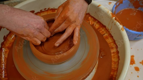The fingers of an artisan potter form an earthen bowl (Close-up). The ceramic product rotates on a potter's wheel. The potter's hands shape the clay bowl from the inside