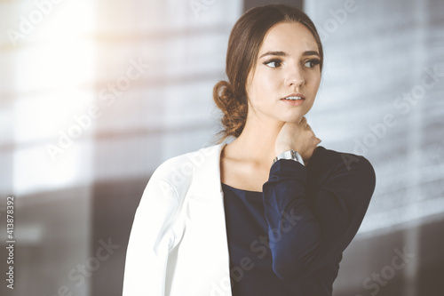 Friendly young business woman or female student dressed in white suit is standing straight and posing at camera in a sunny office. Lifestyle and diverse people concept
