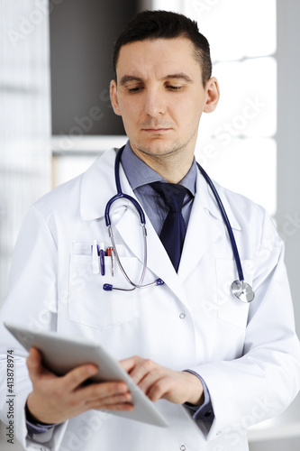 Friendly male doctor using tablet computer in clinic near his working place. Perfect medical service in hospital. Medicine and healthcare concept
