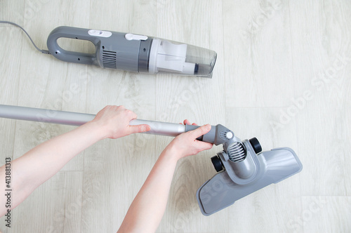 The hand of a Caucasian woman puts a brush attachment on a gray vacuum cleaner on white wooden background.