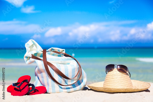 Beach accessories on sand for summer vacation concept. Bag, straw hat with sunglasses and red flip flops. White sand with amazing ocean and blue sky in the background. Free space for your text