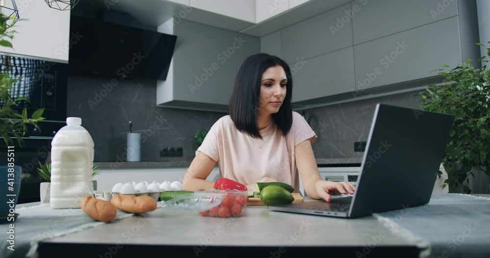 Beautiful smiling satisfied young brunette sitting at kitchen table and watching culinary program online on laptop during preparation vegetable salad for family dinner Beautiful smiling satisfied young brunette sitting at kitchen table and watching culinary program online on laptop during preparation vegetable salad for family dinner