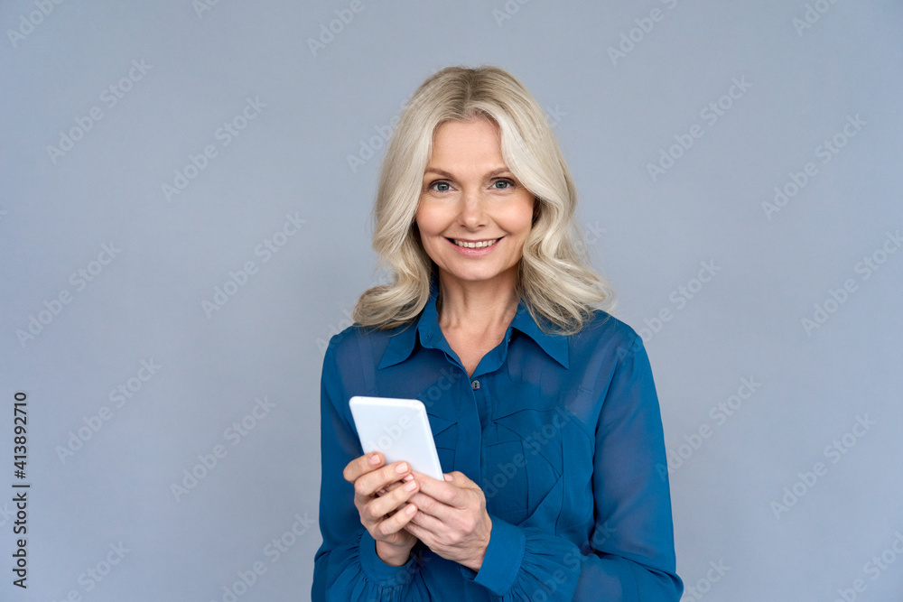 Happy middle aged business woman using apps looking at camera holding smartphone isolated on grey wall. Smiling 50s old lady advertising online mobile e bank services easy applications technology. Happy middle aged business woman using apps looking at camera holding smartphone isolated on grey wall. Smiling 50s old lady advertising online mobile e bank services easy applications technology.