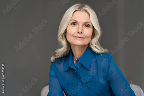 Sophisticated elegant beautiful 50s attractive middle aged smiling woman model sitting in chair looking at camera at home. Portrait of gorgeous confident mature older blond hair lady indoors.