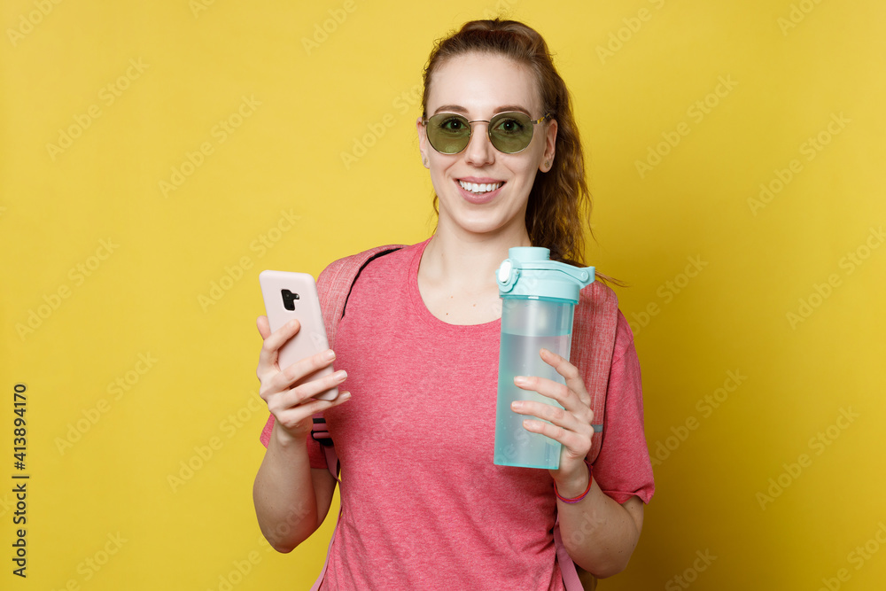 Graceful female standing over yellow background and holding mobile phone and blue bottle of water.