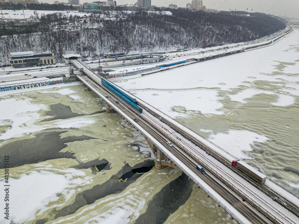 One train on Kiev metro bridge across the frozen Dnieper river. Textured pattern on ice. Aerial drone view. Winter snowy morning.