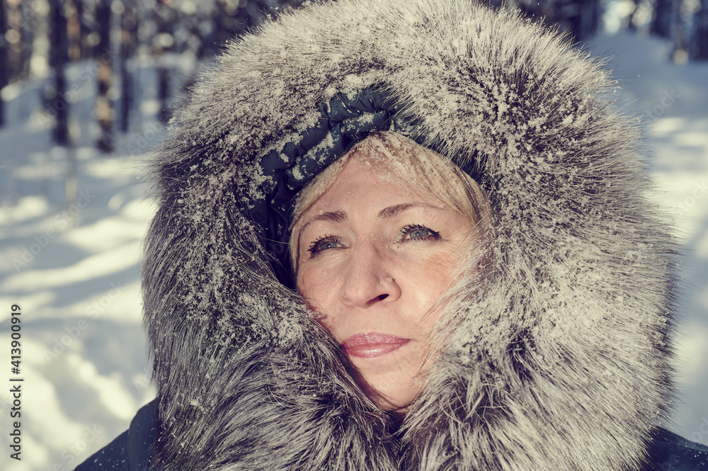 55-60-year-old blonde woman in winter clothes - a jacket with a polar fox edge under sunlight outdoors on the background of a snow-covered forest and snowfall