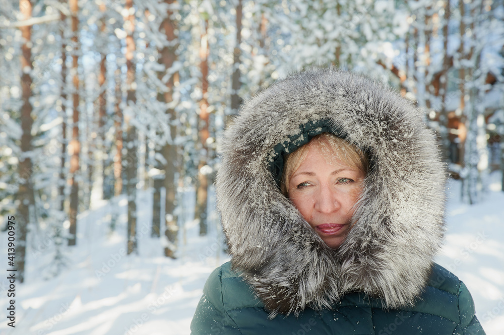 55-60-year-old blonde woman in winter clothes - a jacket with a polar fox edge under sunlight outdoors on the background of a snow-covered forest and snowfall 55-60-year-old blonde woman in winter clothes - a jacket with a polar fox edge under sunlight outdoors on the background of a snow-covered forest and snowfall