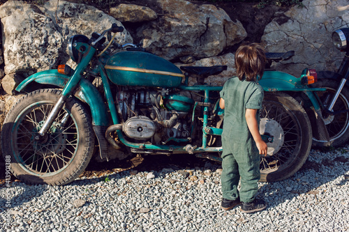 Fototapeta Naklejka Na Ścianę i Meble -  child boy in a green jumpsuit walks among old motorcycles in the summer