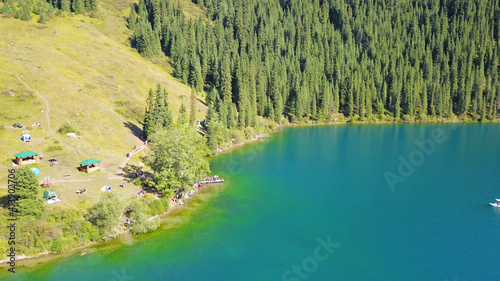 Kolsay lake among green hills and mountains. The mountain lake is surrounded by green forest  tall coniferous trees  grass and bushes. Clean water is like a mirror. Tourists swim on boats. Kazakhstan